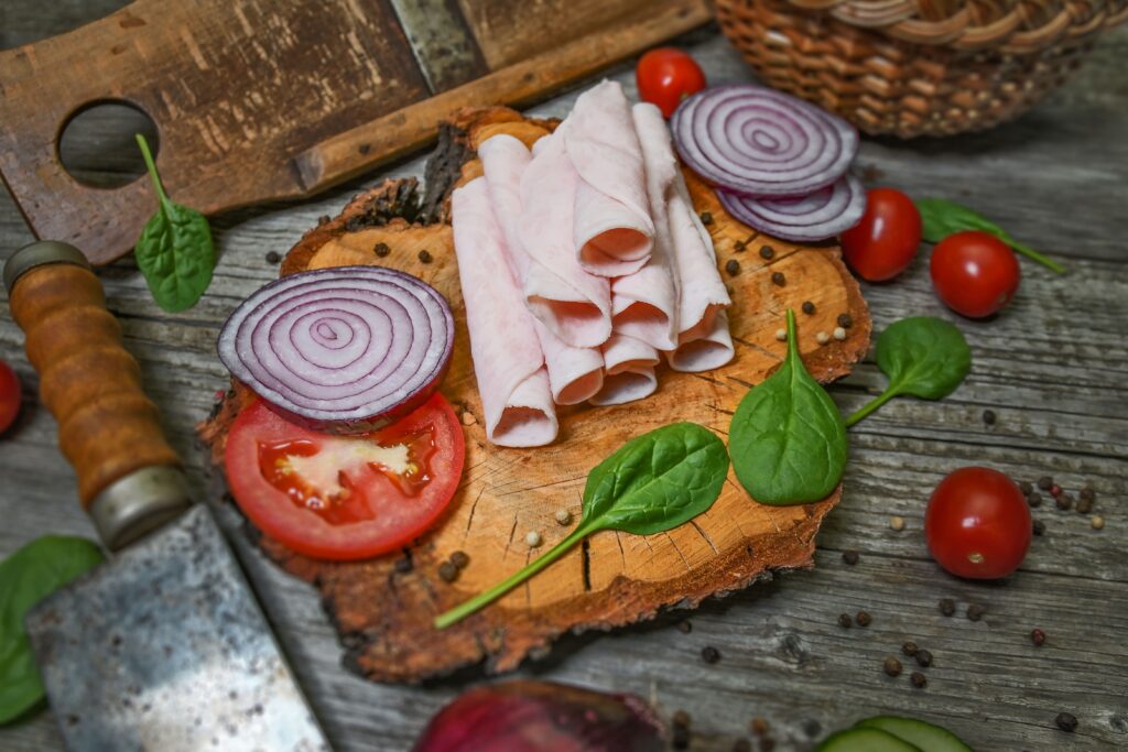 A rustic setup featuring sliced ham, onions, tomatoes, and spinach on a wooden board.